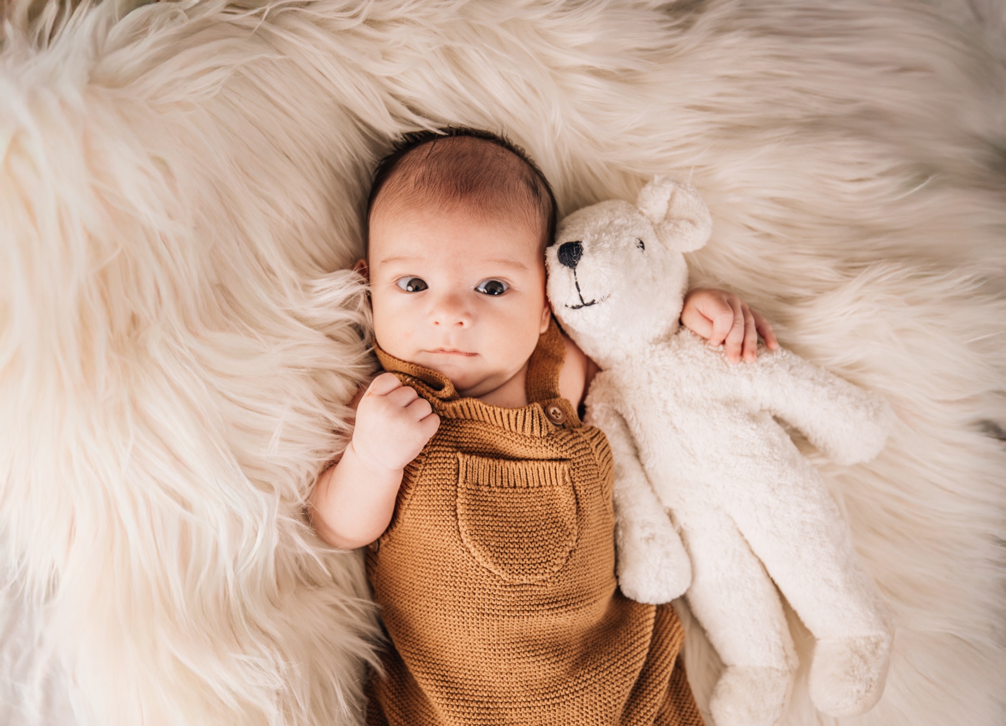 Newborn in a knit romper cuddling a plush bear on a soft fur blanket