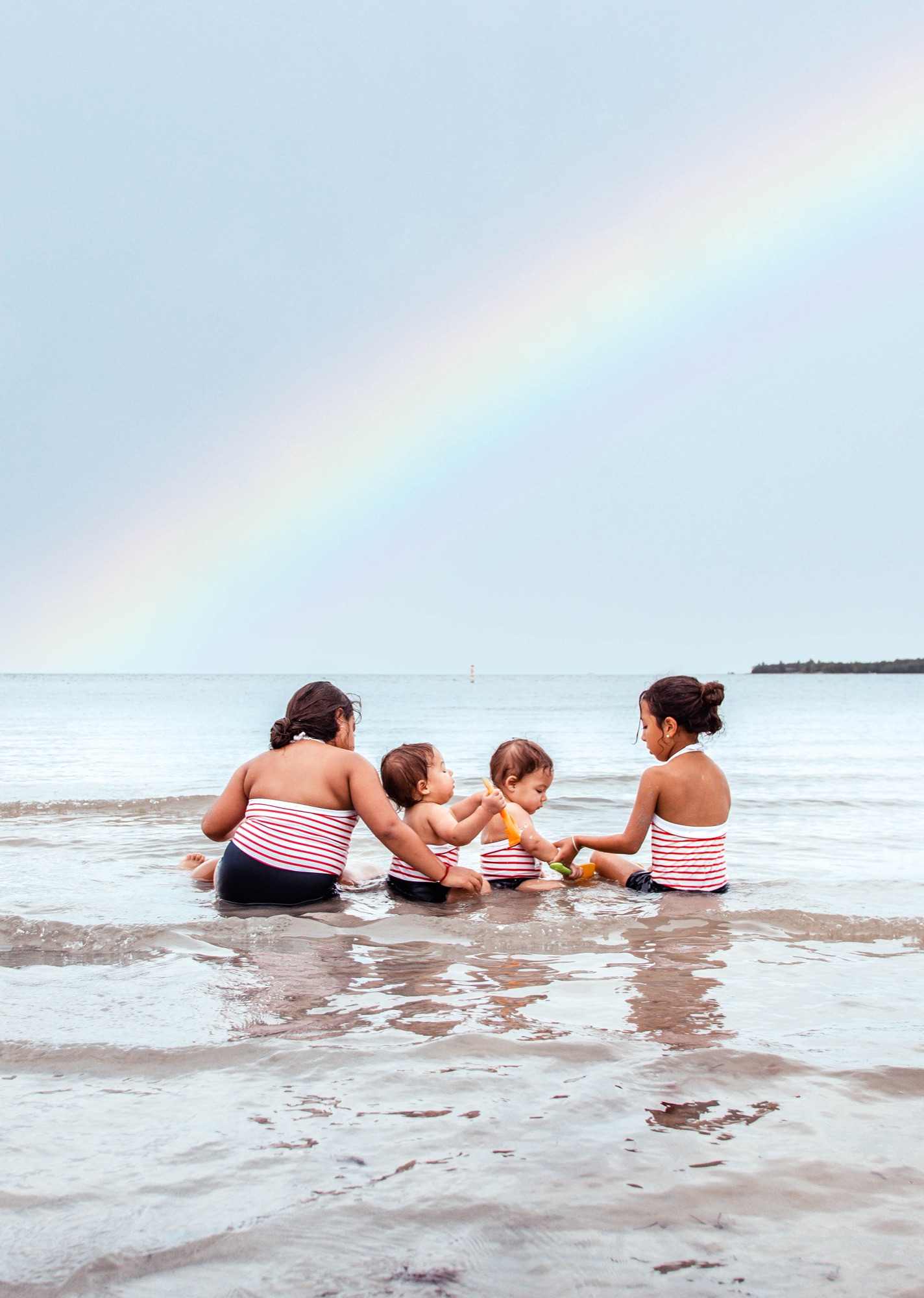 Four sisters in matching striped swimsuits playing in the shallows beneath a rainbow