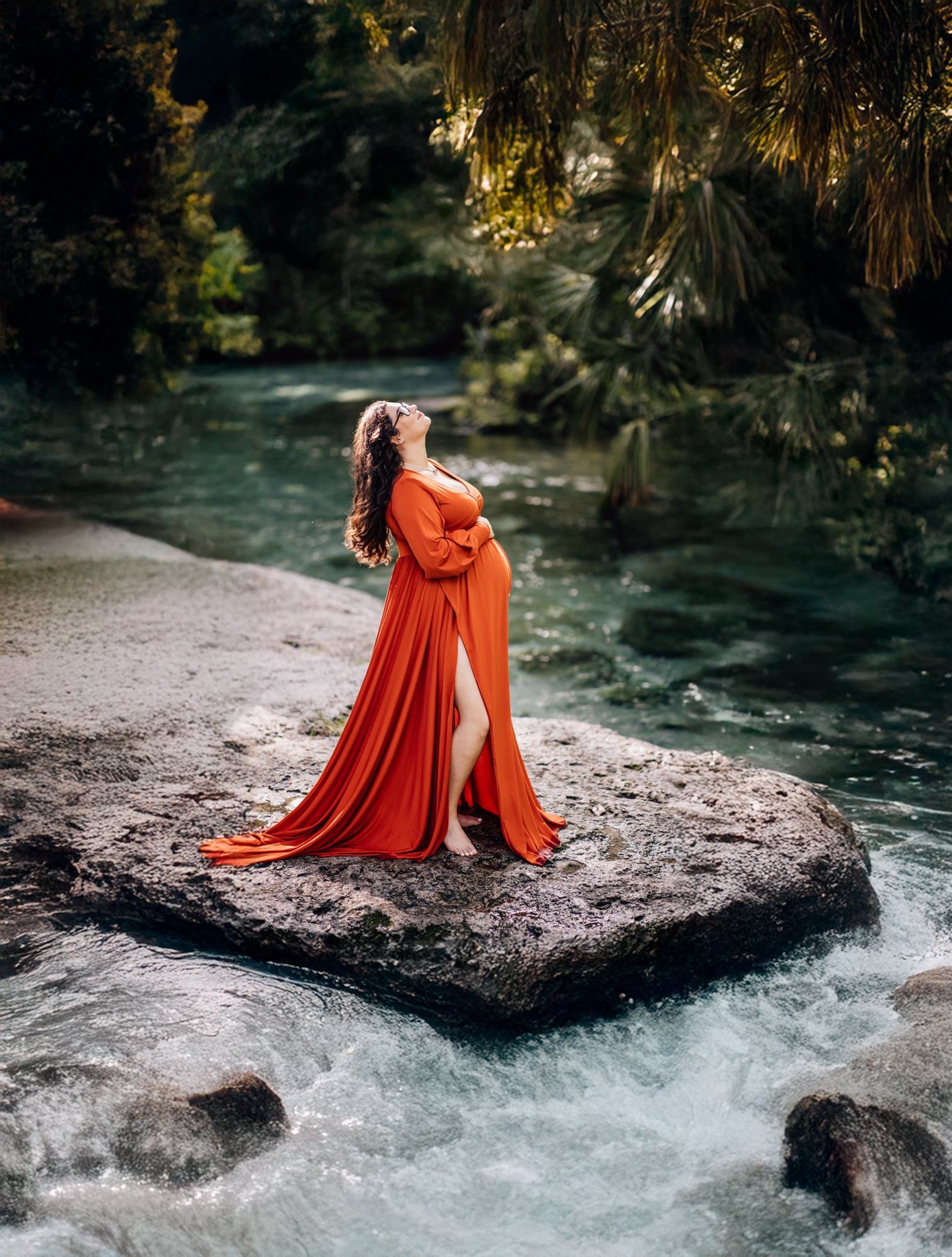 Pregnant woman in a flowing rust orange gown standing on a rock in a turquoise spring surrounded by greenery