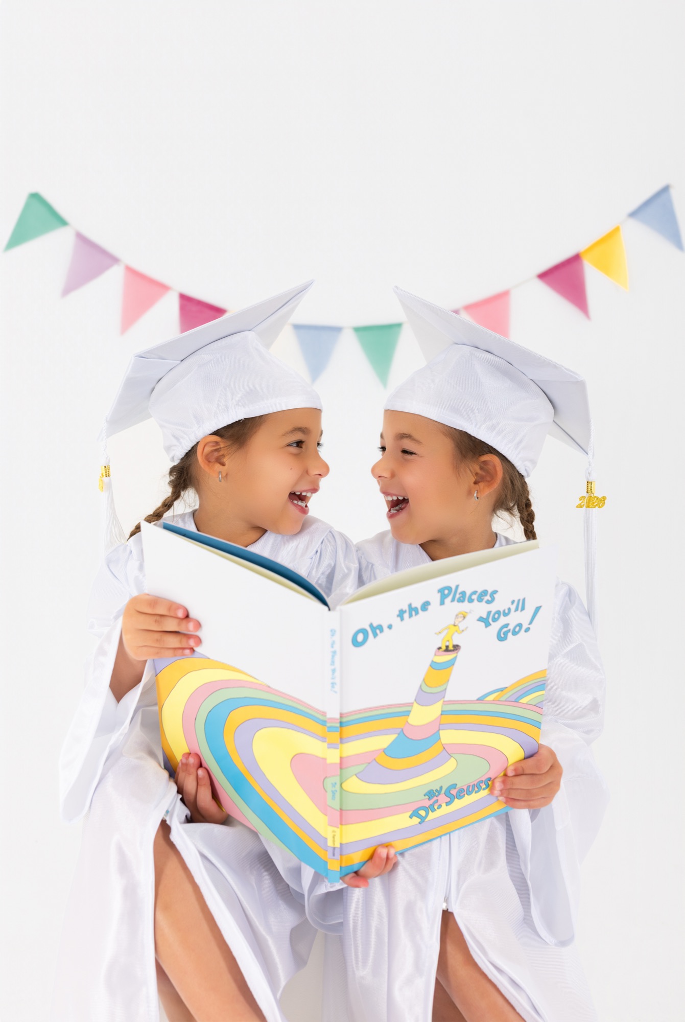 Twin girls in white caps and gowns laughing over a copy of Oh, the Places You'll Go!