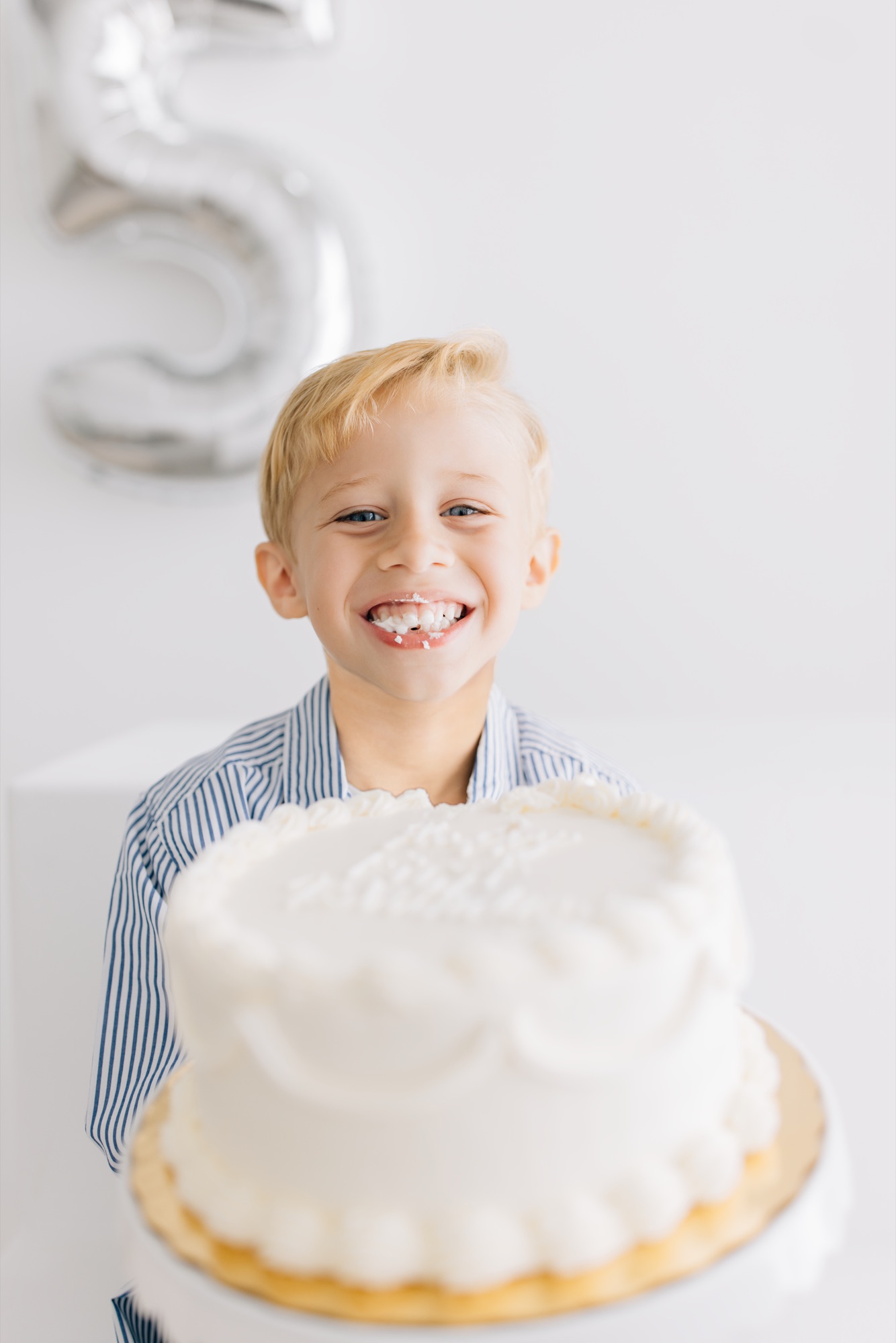 Blond boy grinning with frosting on his mouth behind his fifth birthday cake
