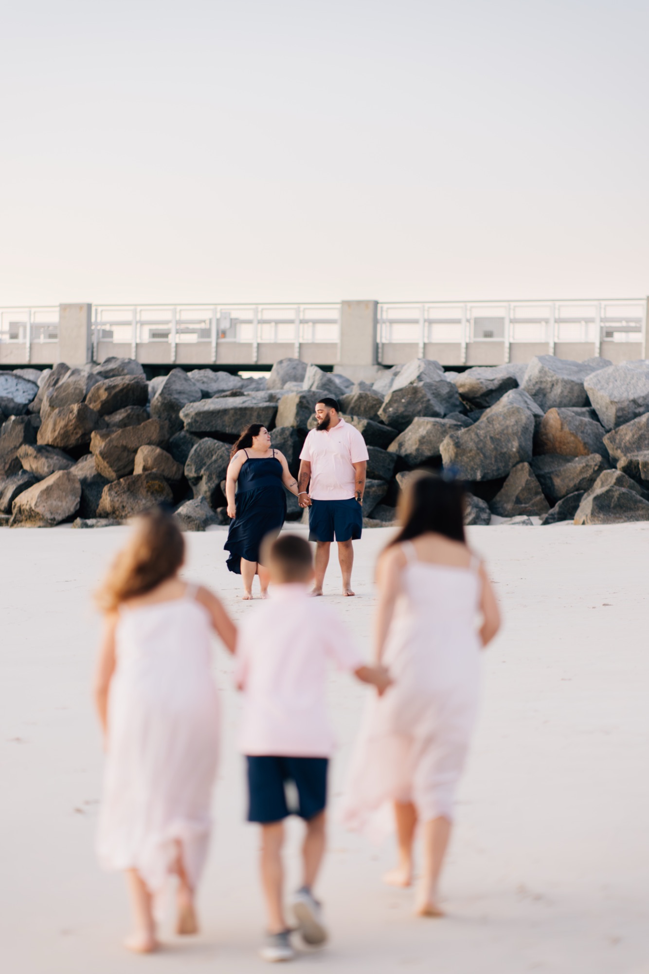 Parents holding hands on a white sand beach at sunset while their three kids run toward them