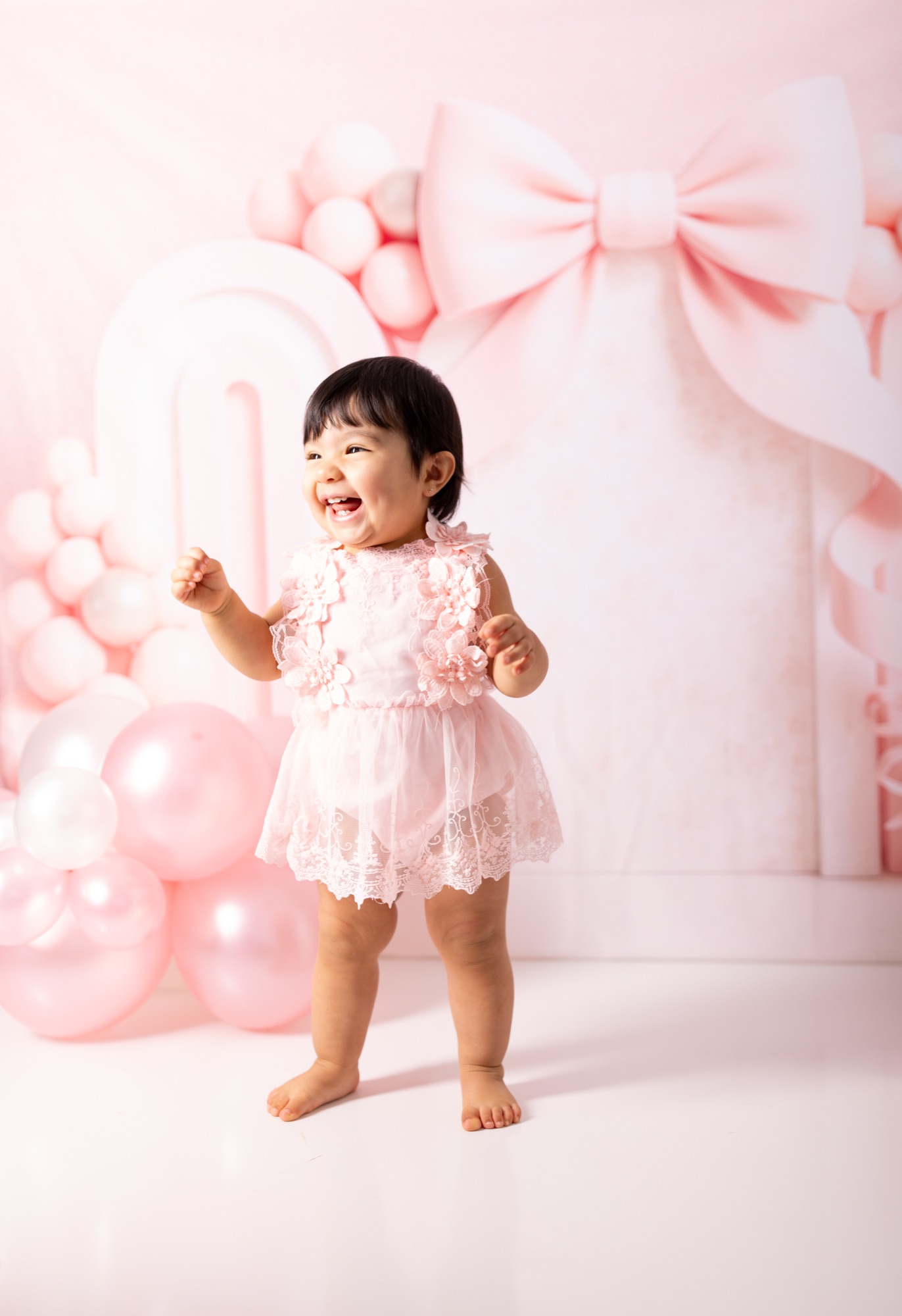 Baby girl in a pink lace romper laughing mid-step at a first birthday studio session