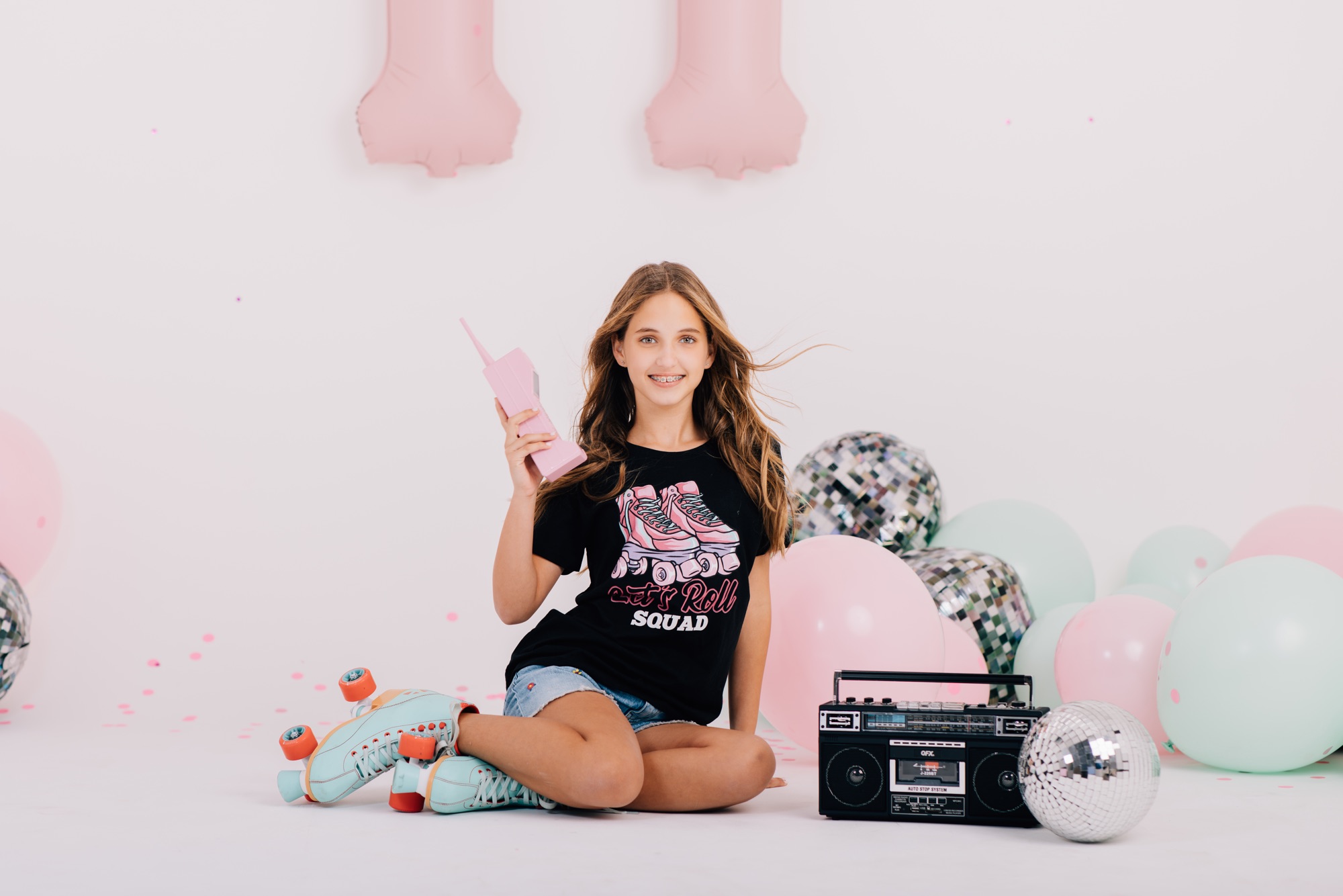 Tween girl posing with a pink retro phone, roller skates, and a disco ball on a white studio set