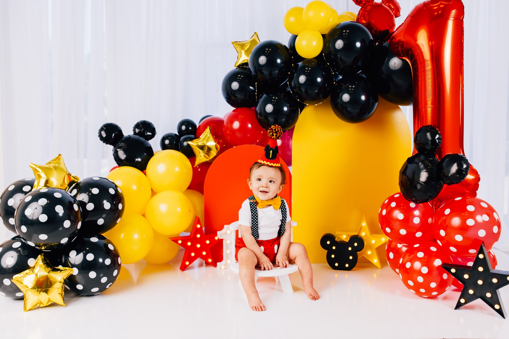 Baby boy in a party hat sitting on a white stool in front of a red and yellow Mickey balloon set