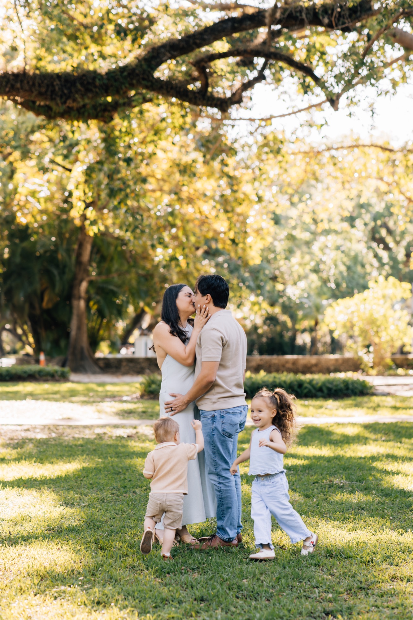 Parents kissing under a large oak while their toddler boy and girl stand at their legs at golden hour