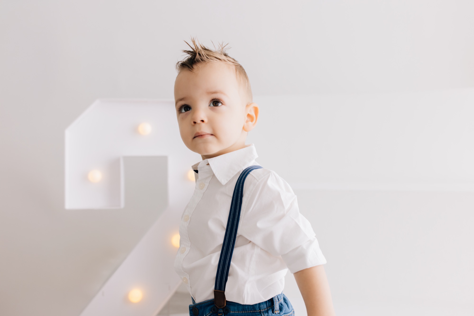 Toddler boy in a white shirt and navy suspenders beside a lit number 2 marquee