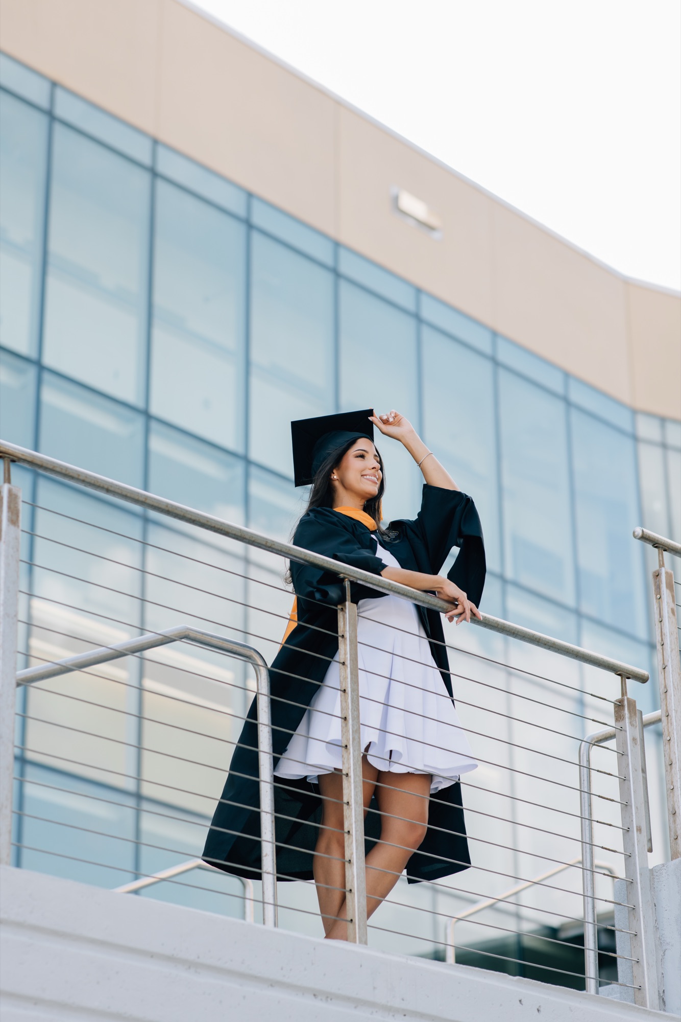 Graduate in cap and gown with a gold stole outside a glass campus building
