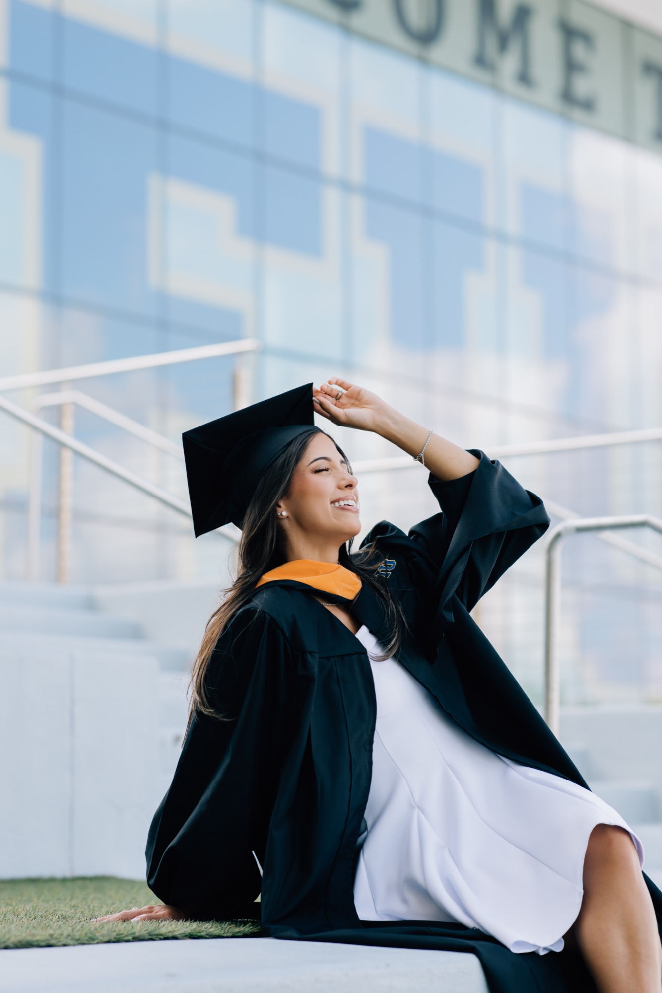Graduate seated on campus steps laughing with a hand on her cap