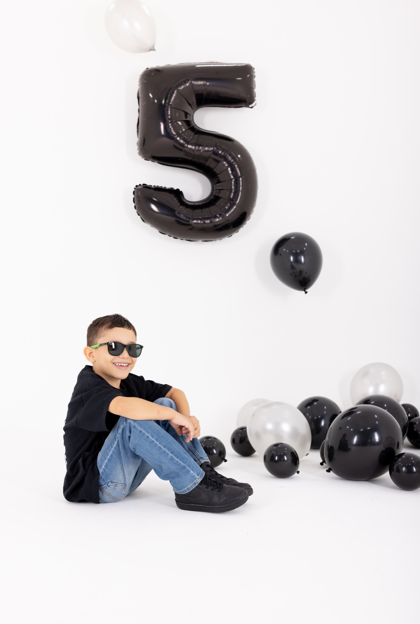 Boy in sunglasses standing under a black number 5 balloon surrounded by black and silver balloons
