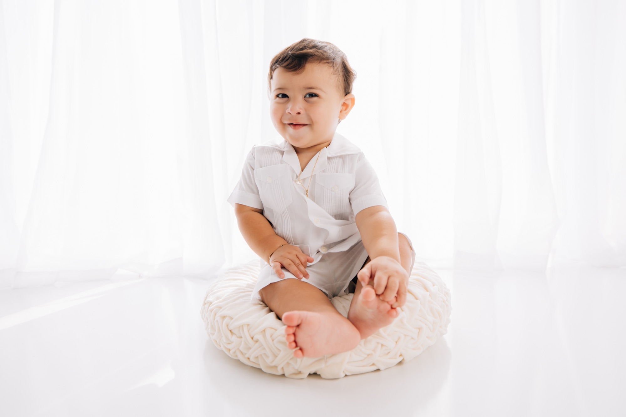 Smiling baby boy in a white guayabera on a cream cushion