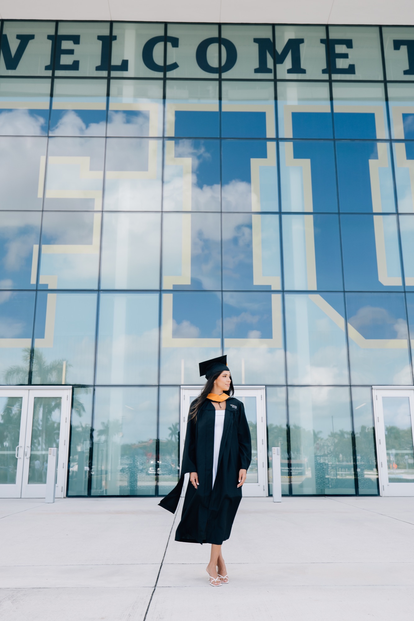 Graduate in a black cap and gown with a gold stole outside FIU's glass building