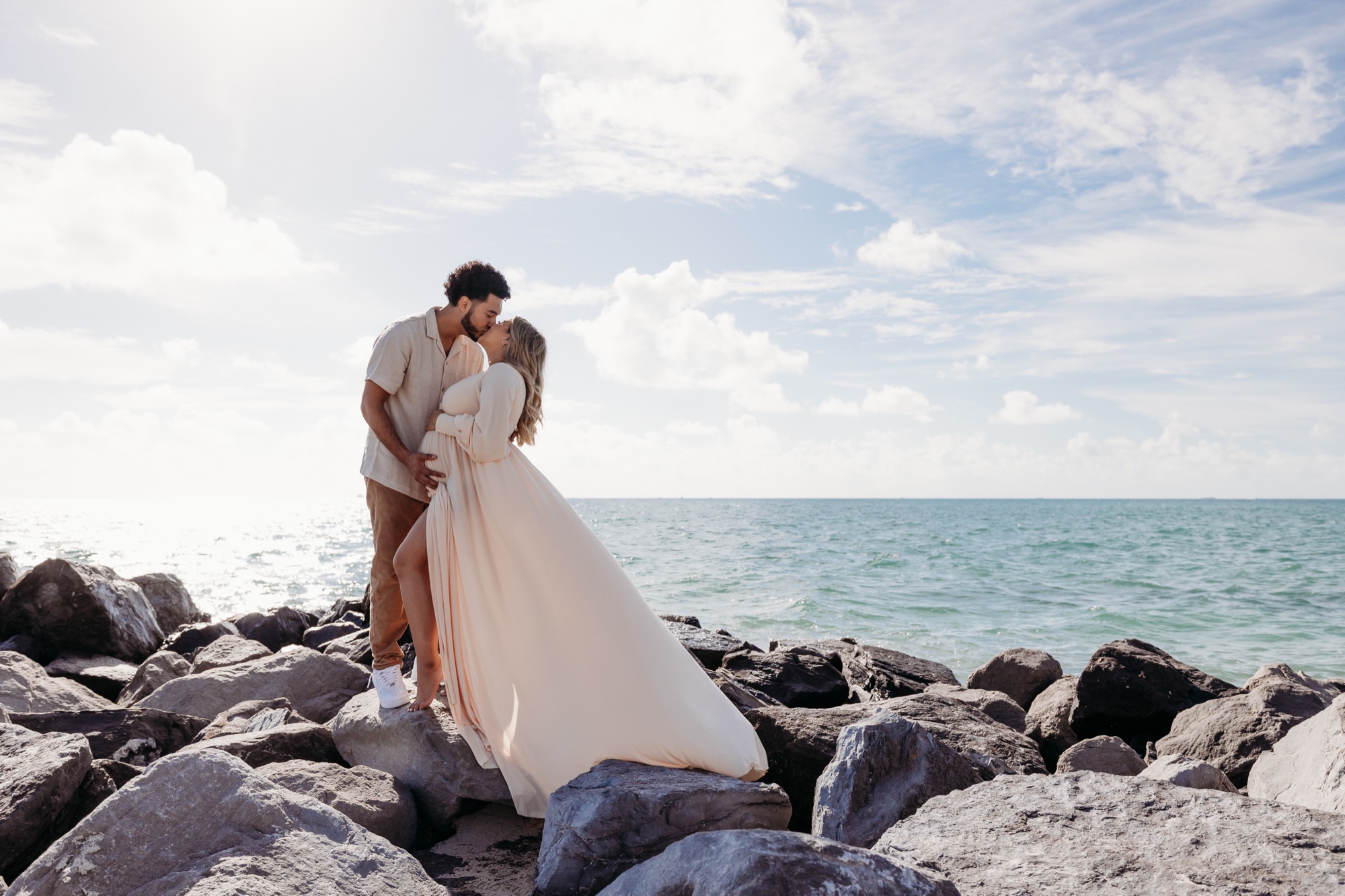 Expectant couple kissing on jetty rocks over teal ocean, the mother in a cream chiffon gown