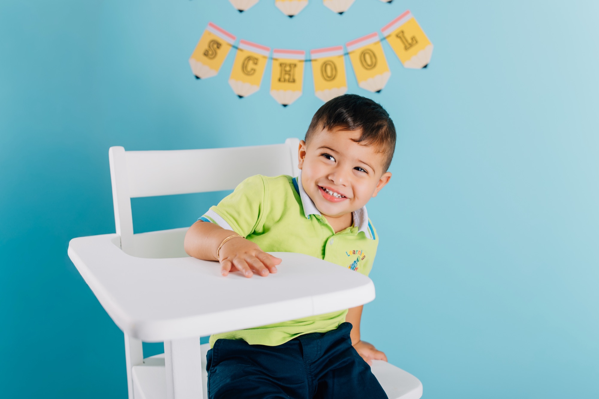 Toddler boy in a green polo grinning at a white school desk in front of an aqua backdrop with a pencil school banner