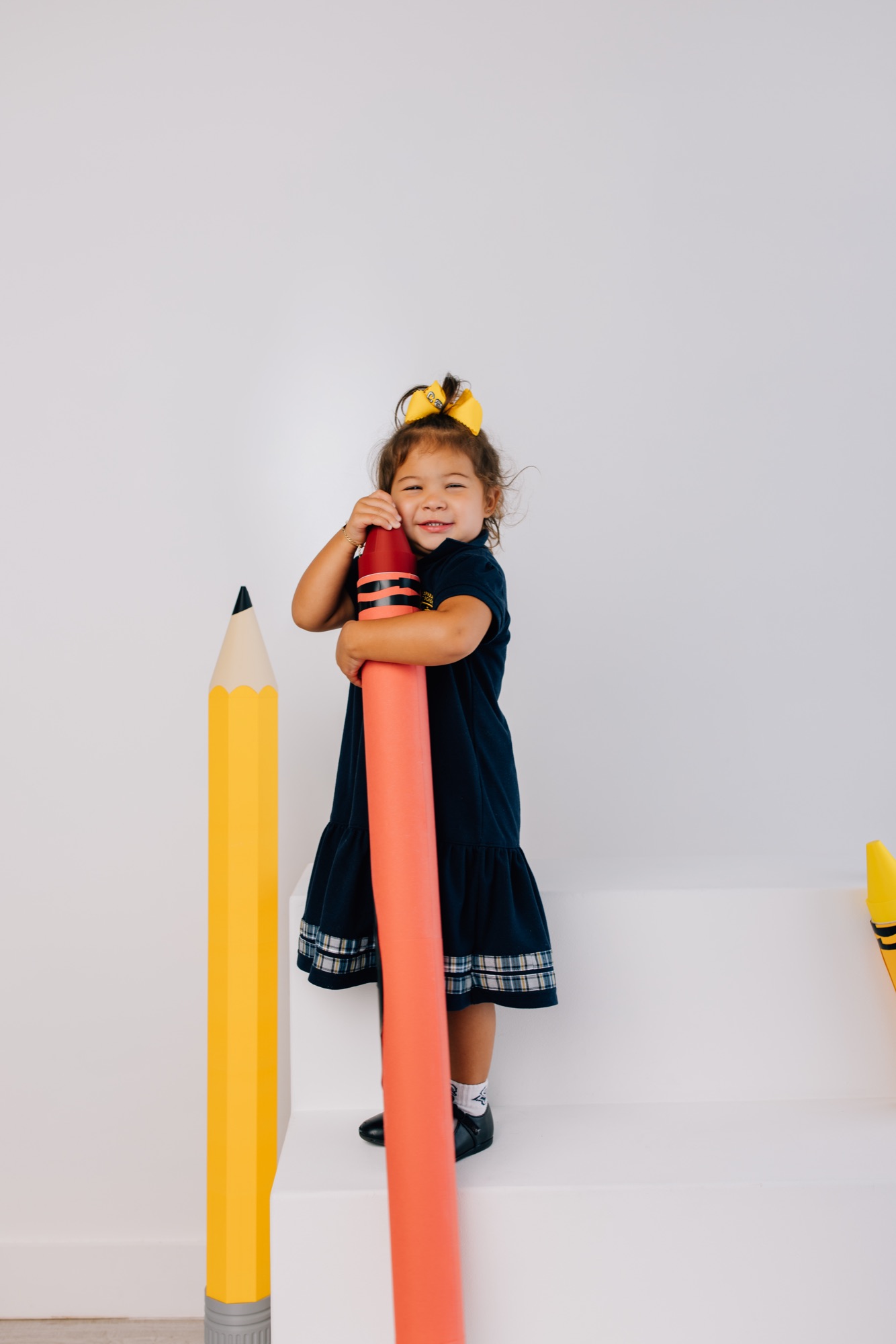 Girl in a navy school uniform with a yellow bow hugging a giant red crayon prop