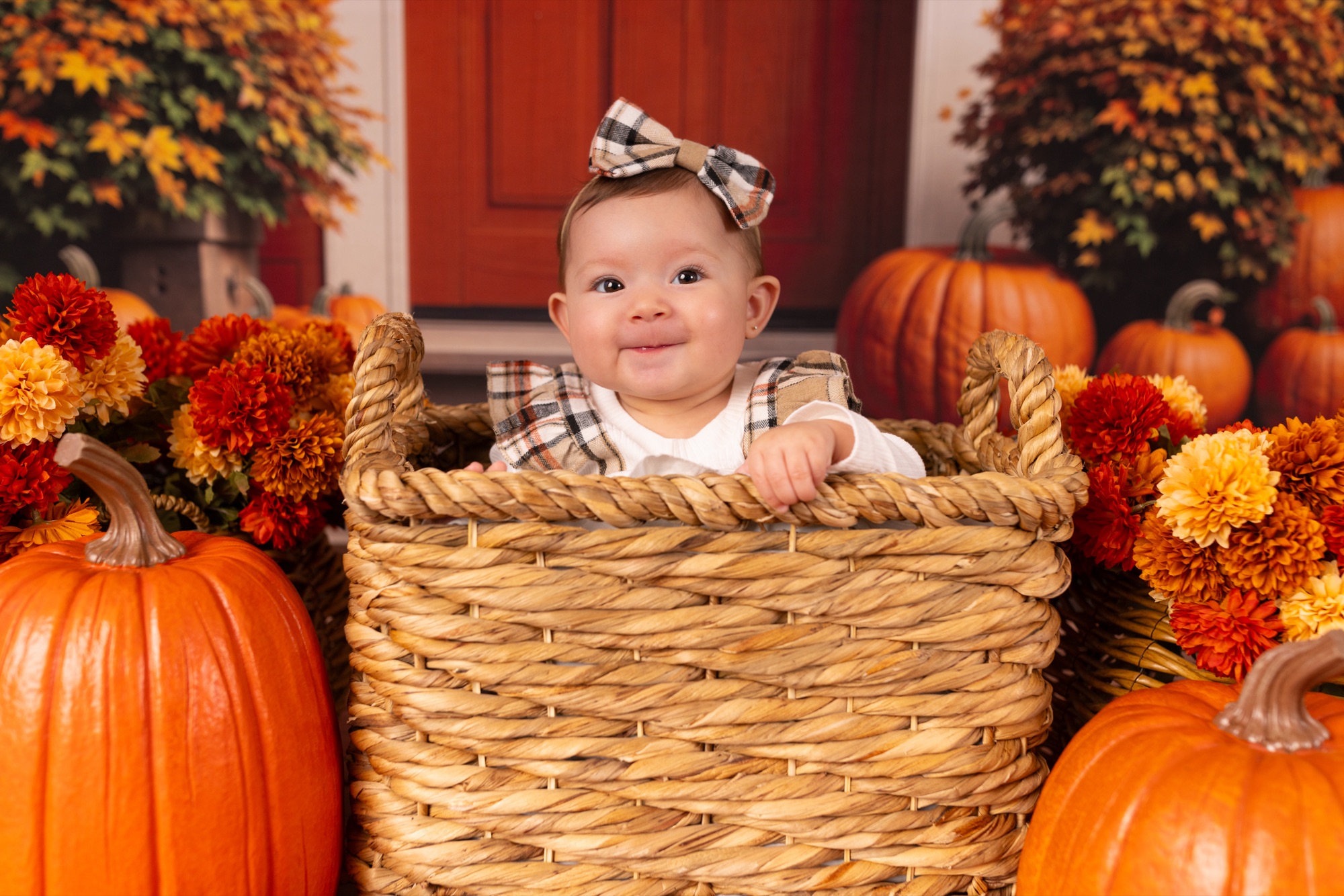 Smiling baby girl with a plaid bow peeking from a woven basket among pumpkins