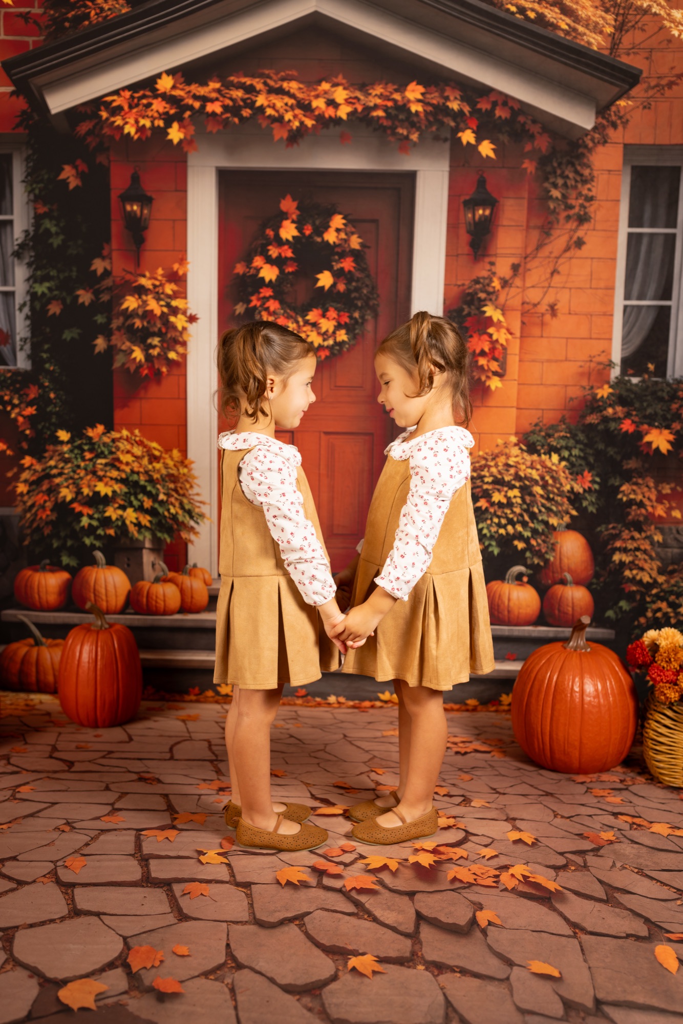 Twin girls in matching tan pinafores holding hands in front of an autumn front door backdrop