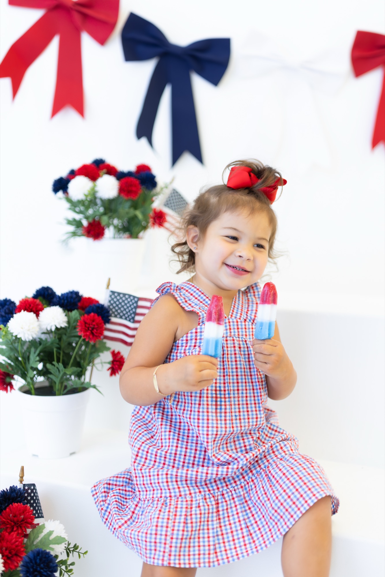 Toddler girl in a red, white, and blue gingham dress holding two rocket popsicles