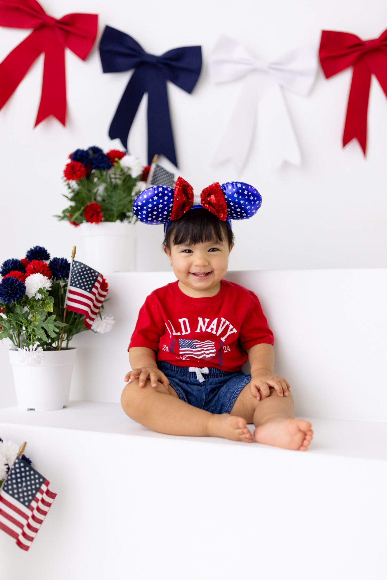 Baby girl in a flag tee and sequined patriotic bow sitting on white steps
