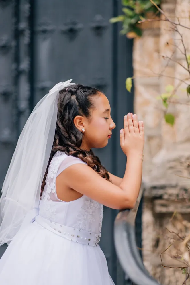 First communion portrait, hands clasped in prayer beside a weathered iron door
