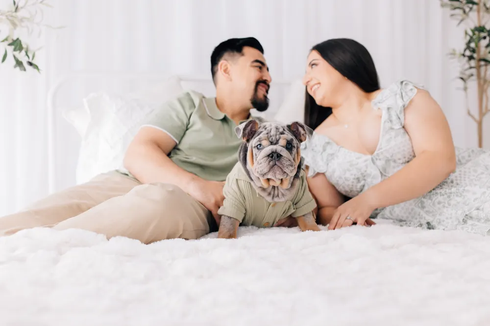 Couple on a white bed smiling at each other with their English bulldog puppy front and center
