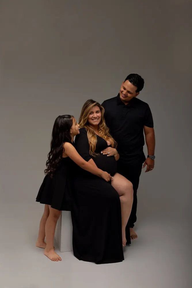 Pregnant mom seated with her daughter leaning on her and dad standing behind, matte grey studio backdrop