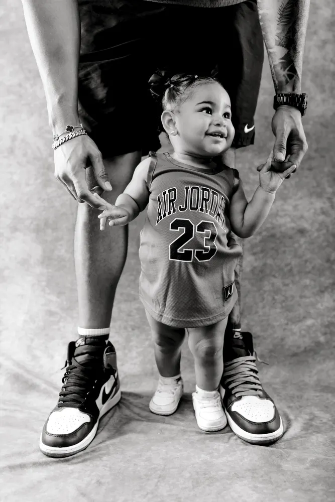 Black and white portrait of a toddler in a Jordan jersey holding her dad’s hand, both in matching sneakers