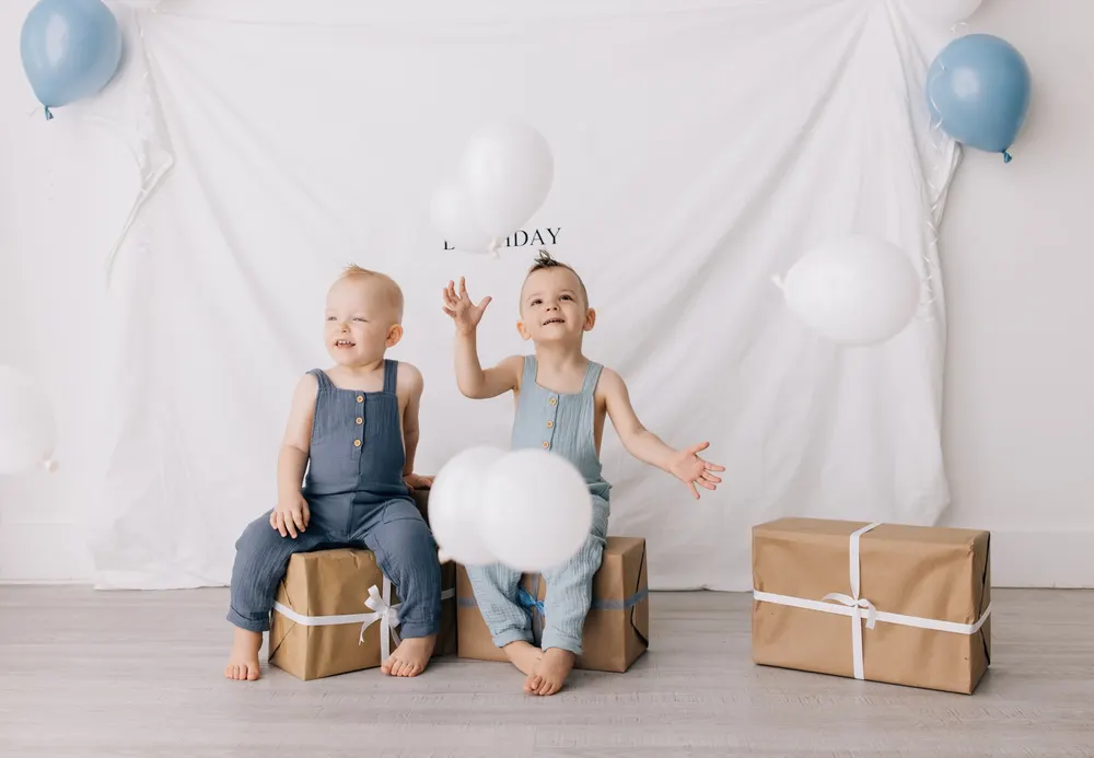Toddler brothers seated on kraft paper presents as white balloons fall around them