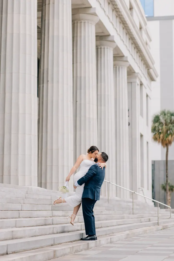 Groom in a navy suit lifting and kissing his bride on courthouse steps framed by stone columns and a palm tree