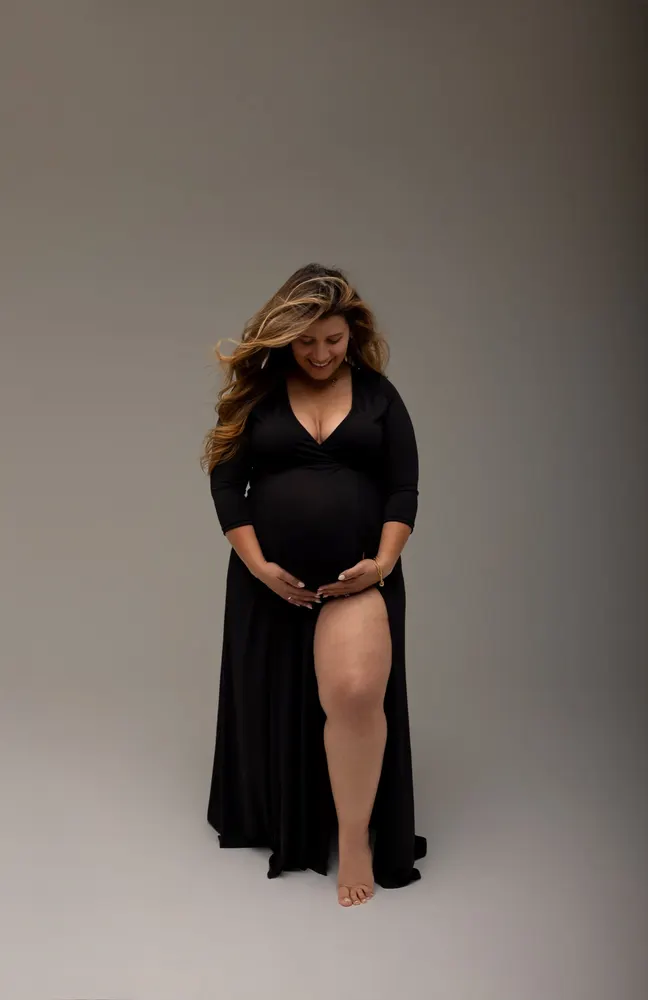 Pregnant woman in a black gown smiling down at her hands on her bump against a grey studio backdrop
