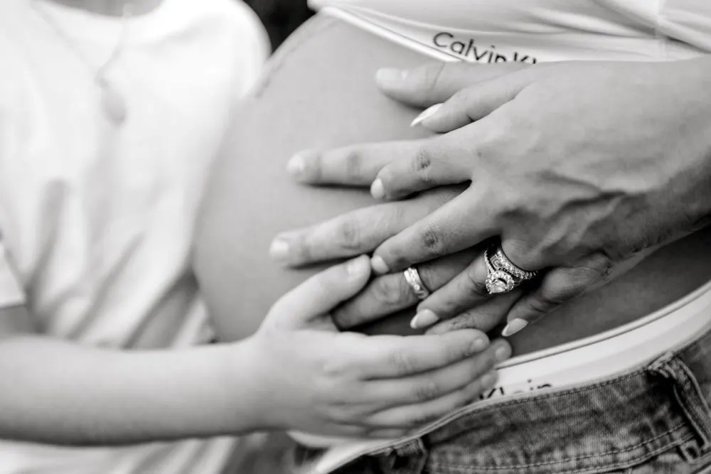 Black-and-white close-up of hands resting on a pregnant belly, wedding rings on display