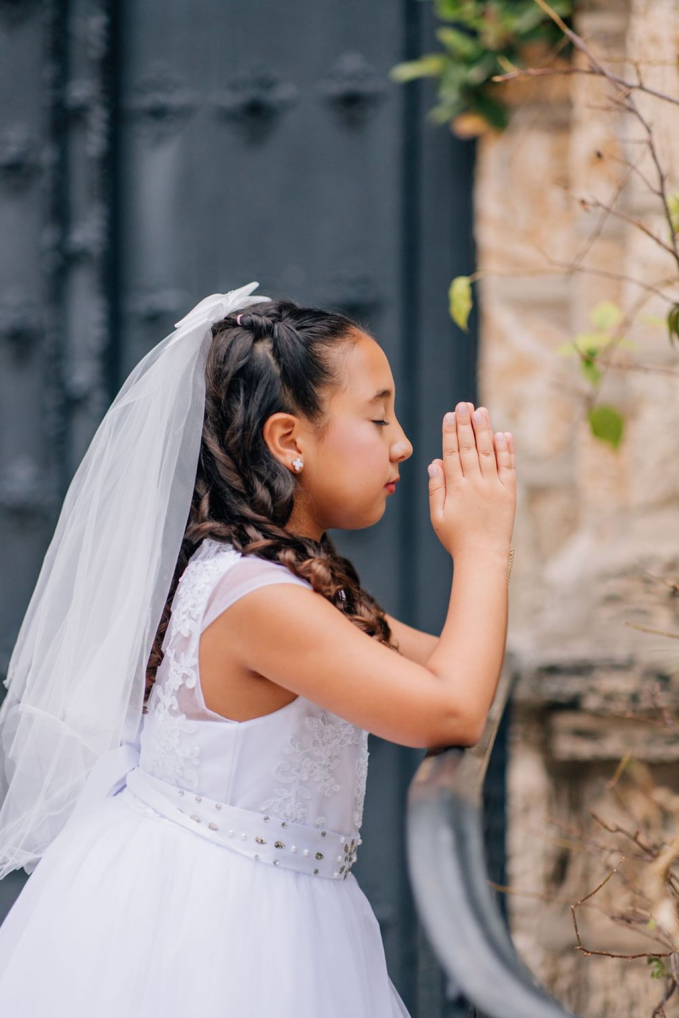 First communion portrait, hands clasped in prayer beside a weathered iron door