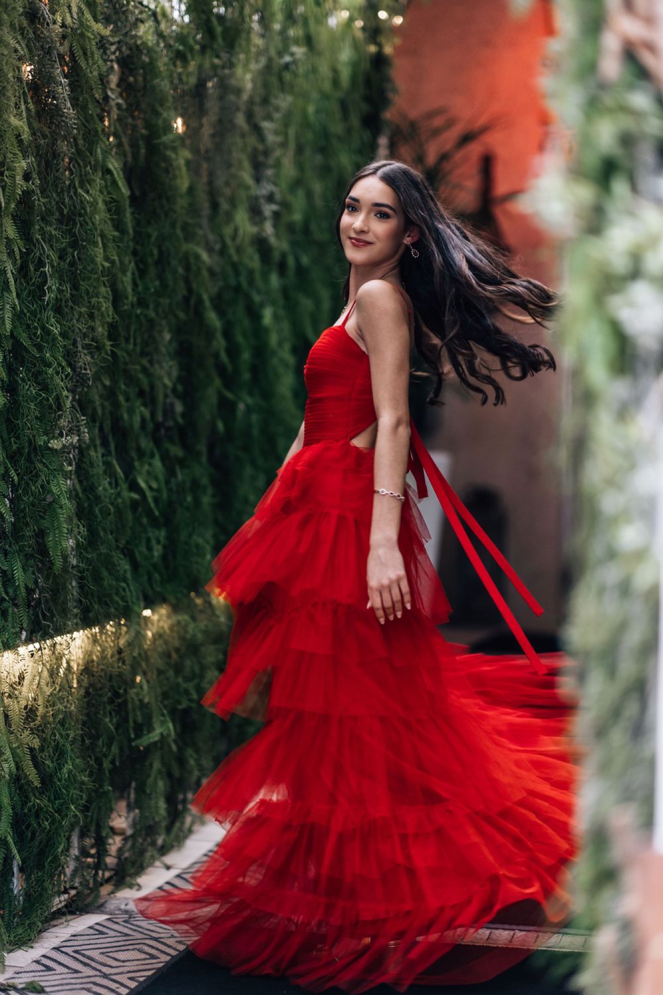 Woman twirling in a red tulle gown through a garden corridor