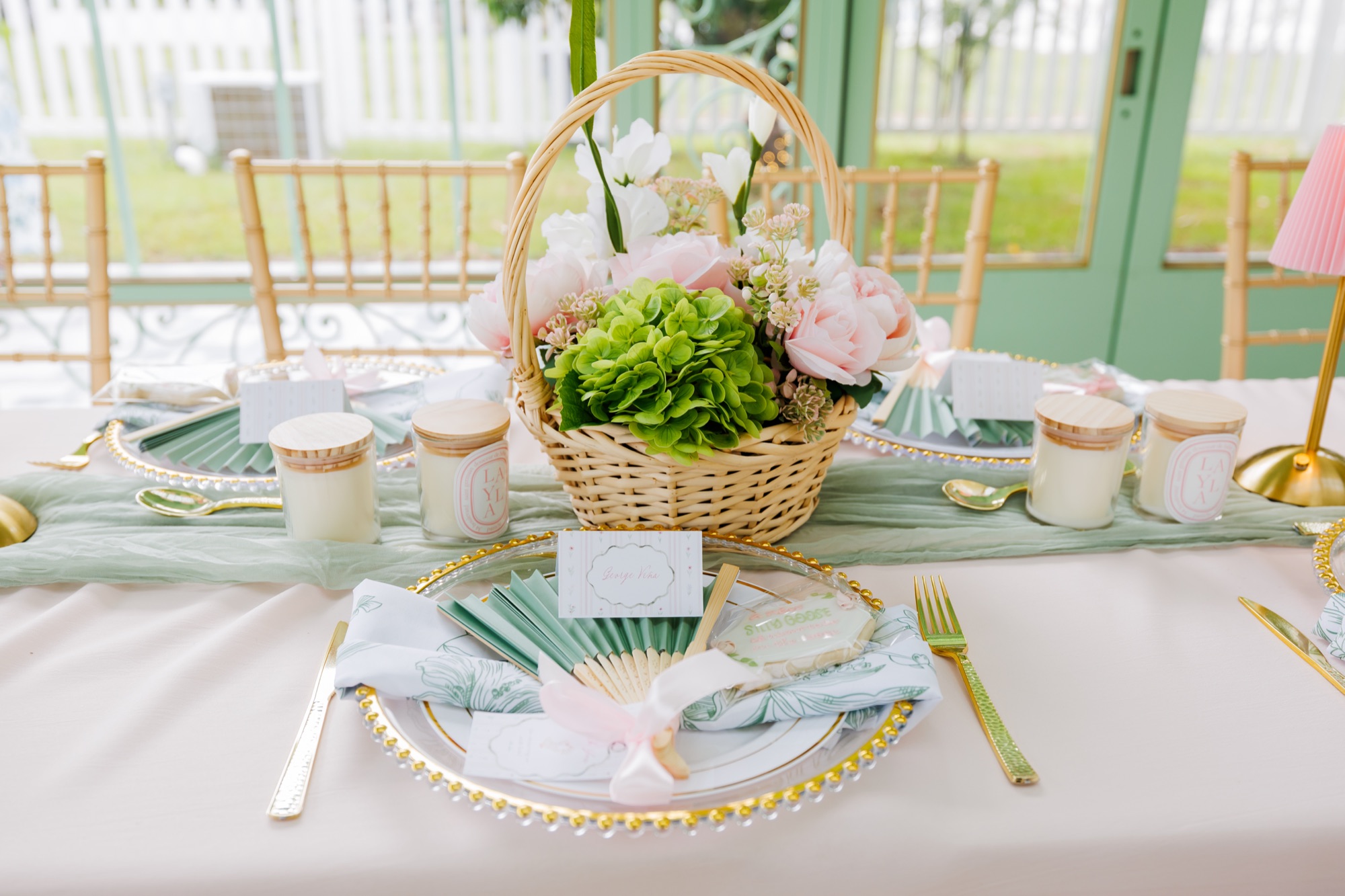 Pastel party table with a wicker flower basket centerpiece and gold flatware in a greenhouse venue