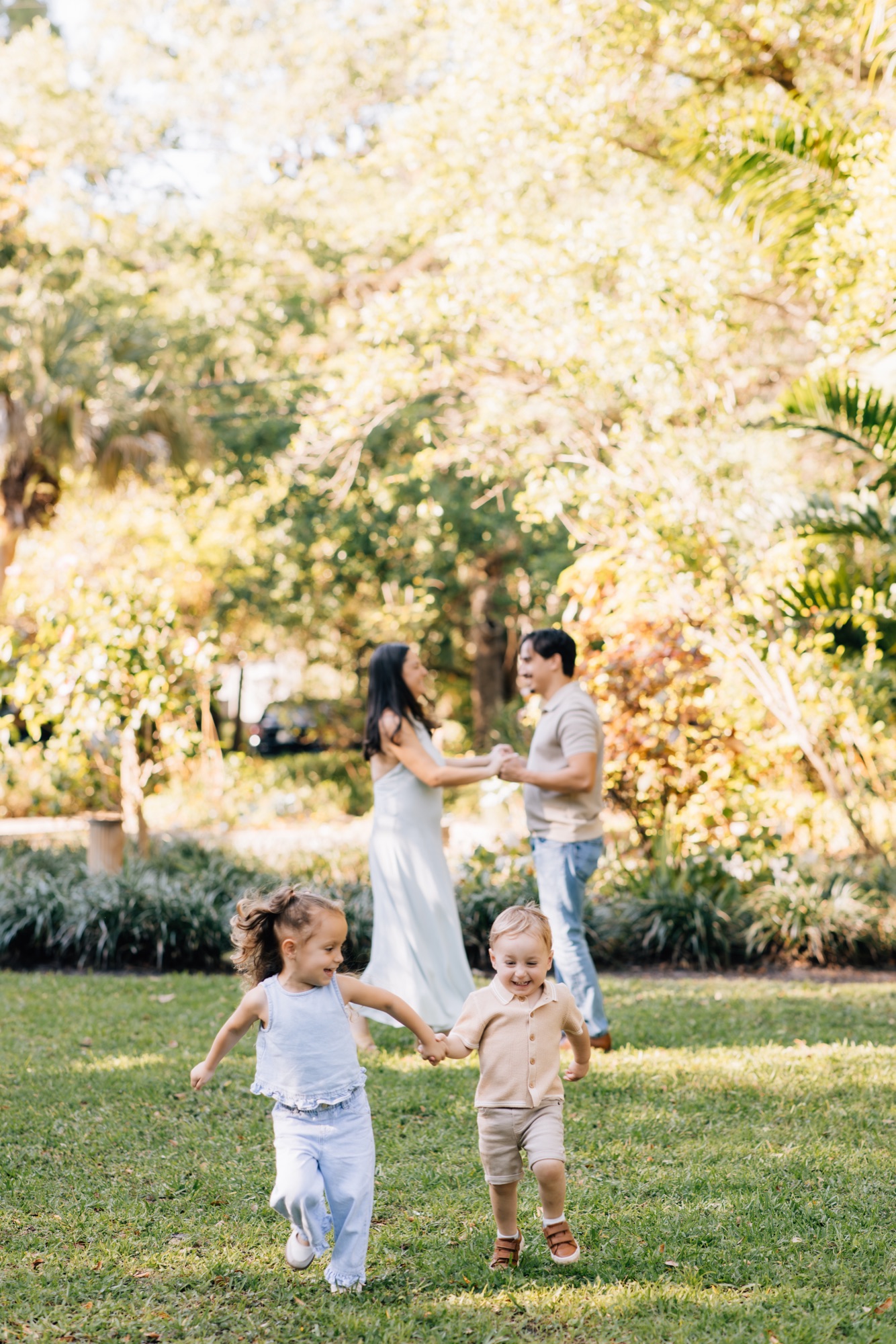 Toddler brother and sister running hand in hand while their parents dance behind them in a sunlit park