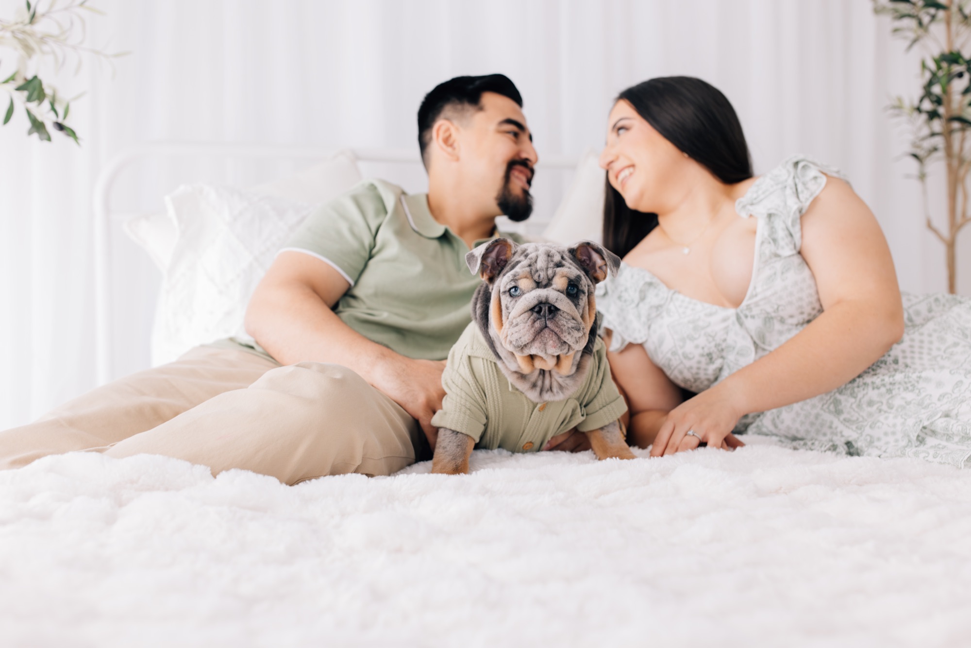 Couple on a white bed smiling at each other with their English bulldog puppy front and center