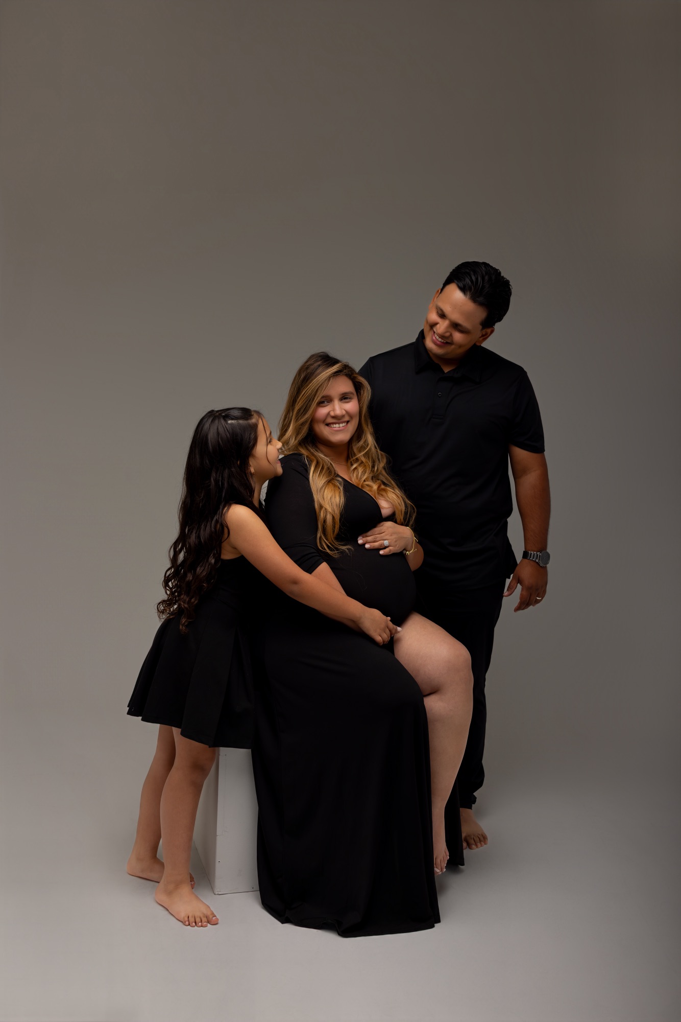 Pregnant mom seated with her daughter leaning on her and dad standing behind, matte grey studio backdrop