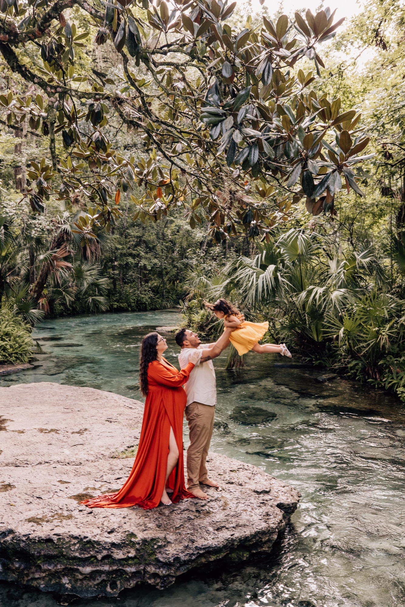 Family on a limestone rock over a clear spring, mom in a flowing red dress and dad lifting their toddler