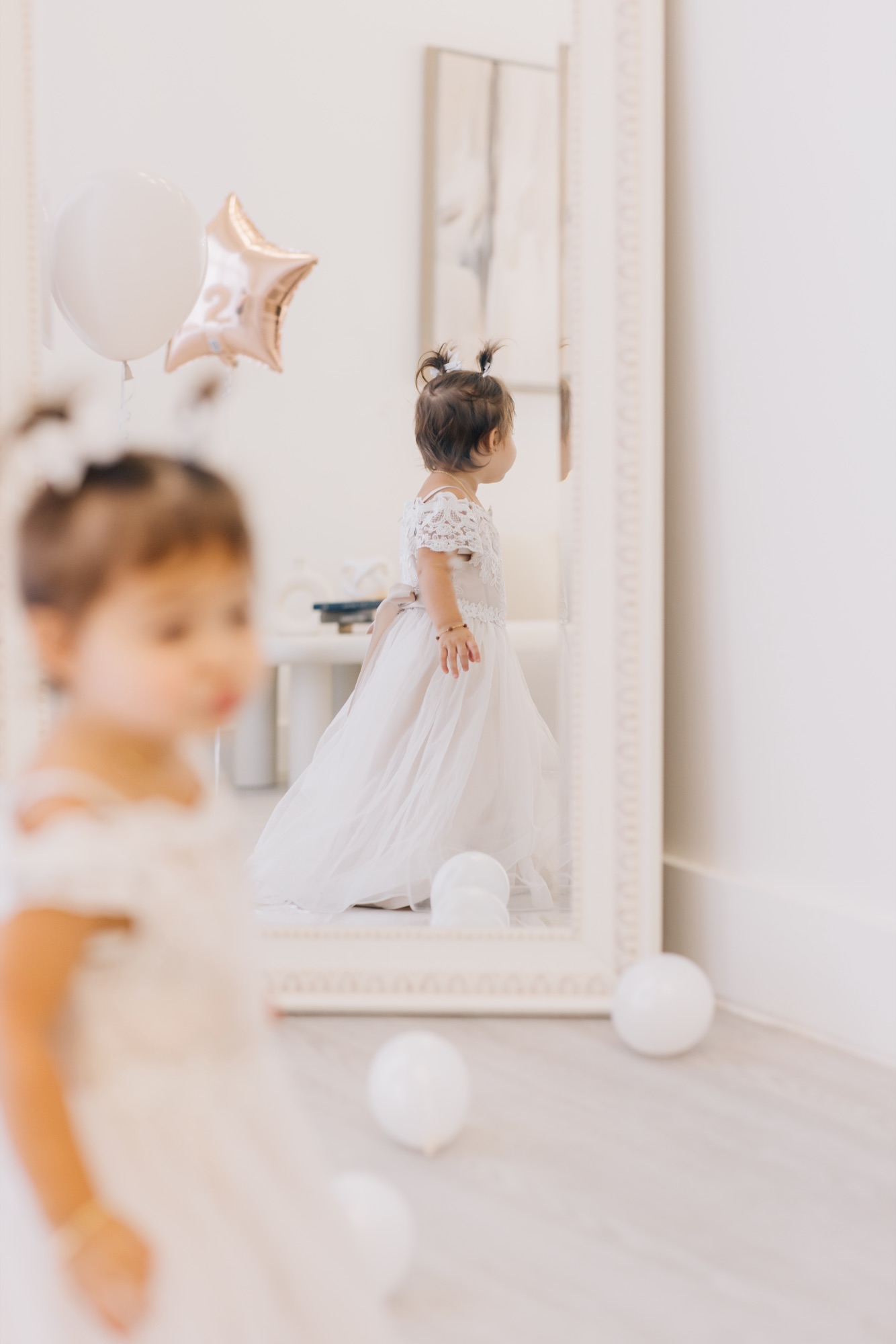 Toddler in a white lace dress reflected in a tall mirror beside a star-shaped number 2 balloon