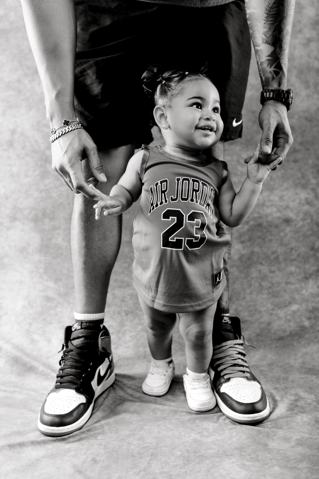 Black and white portrait of a toddler in a Jordan jersey holding her dad’s hand, both in matching sneakers
