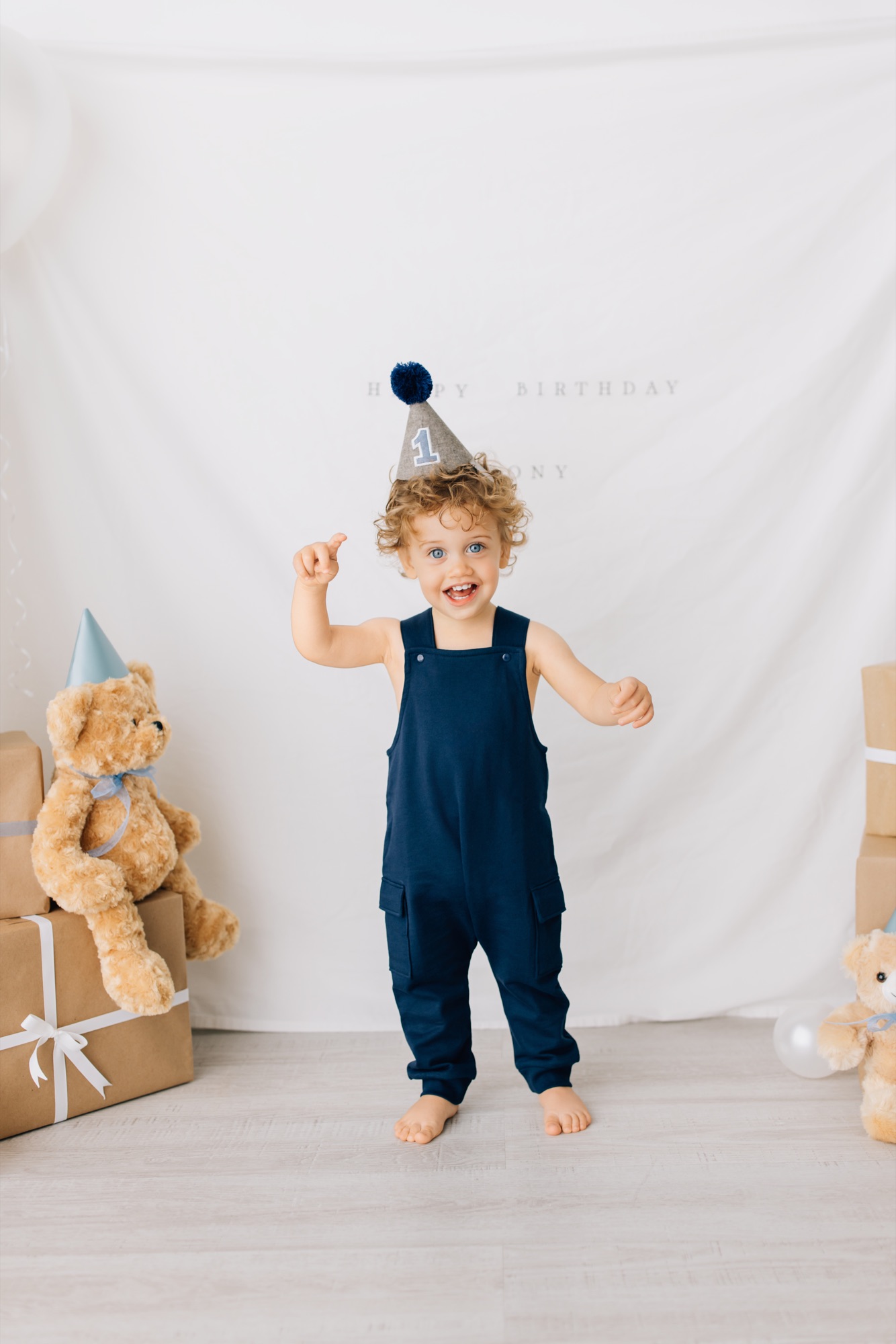 Baby boy in navy overalls and a party hat surrounded by teddy bears at his first birthday session