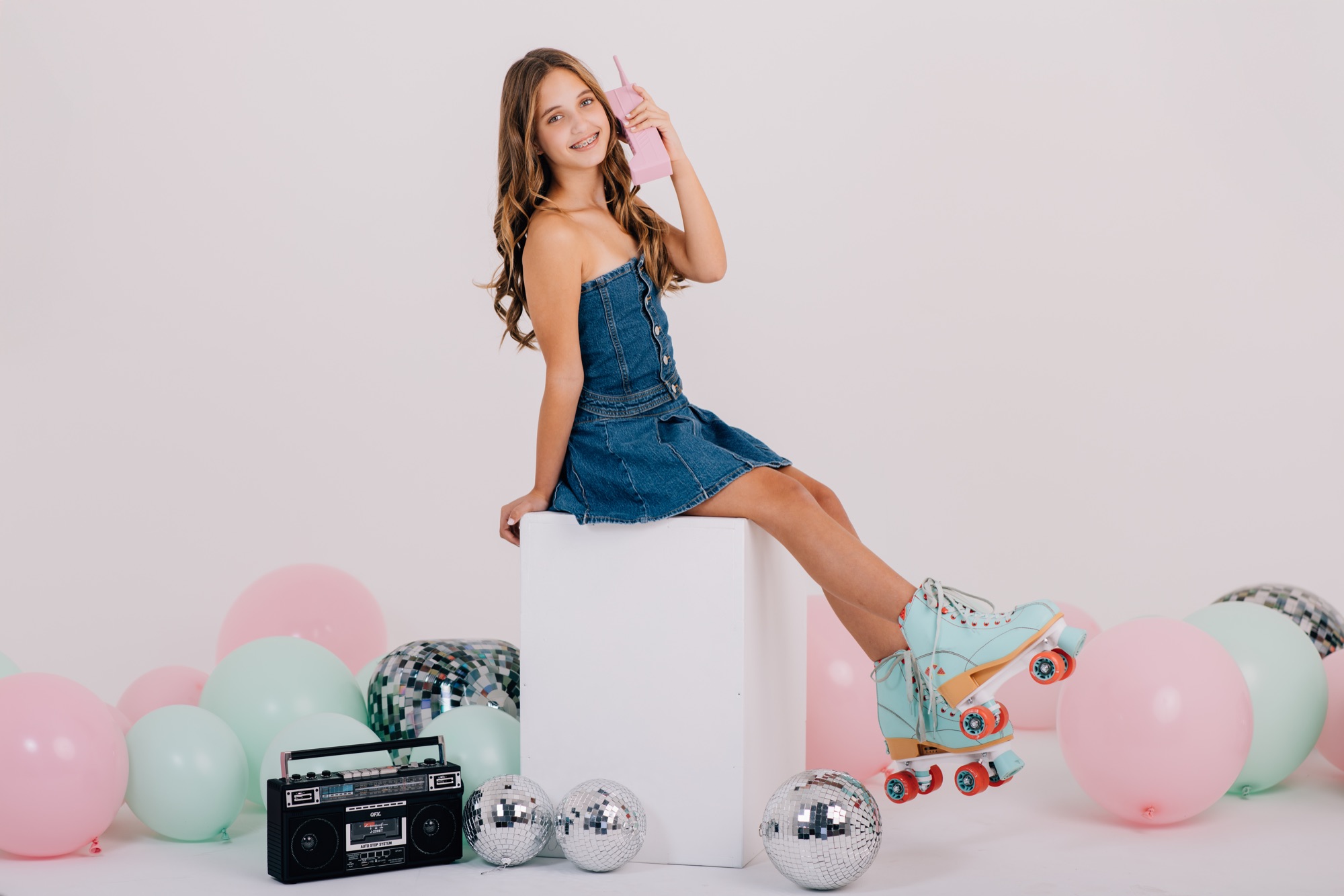 Girl seated on a white cube holding a pink retro phone, roller skates up, surrounded by a pastel skate set