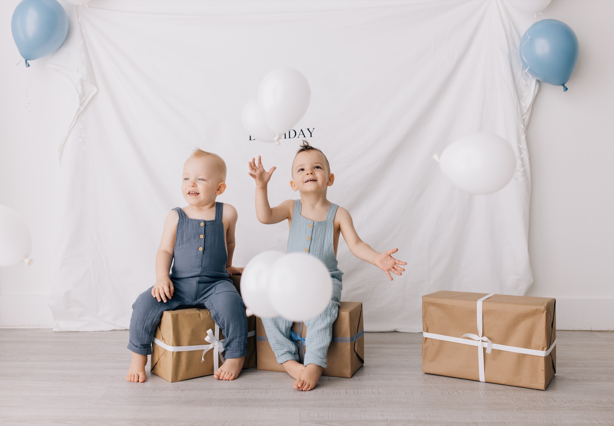 Toddler brothers seated on kraft paper presents as white balloons fall around them