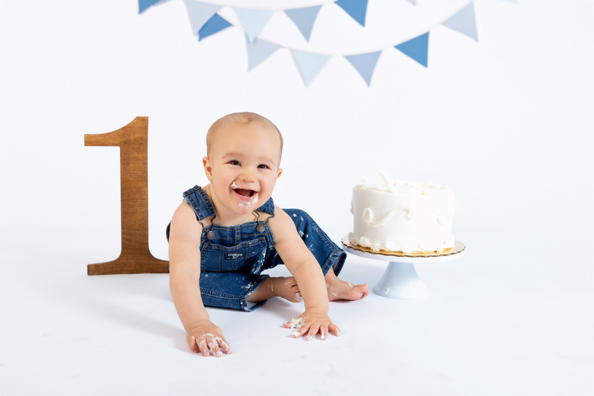 Baby boy in denim overalls grinning with frosting on his face behind his first birthday cake