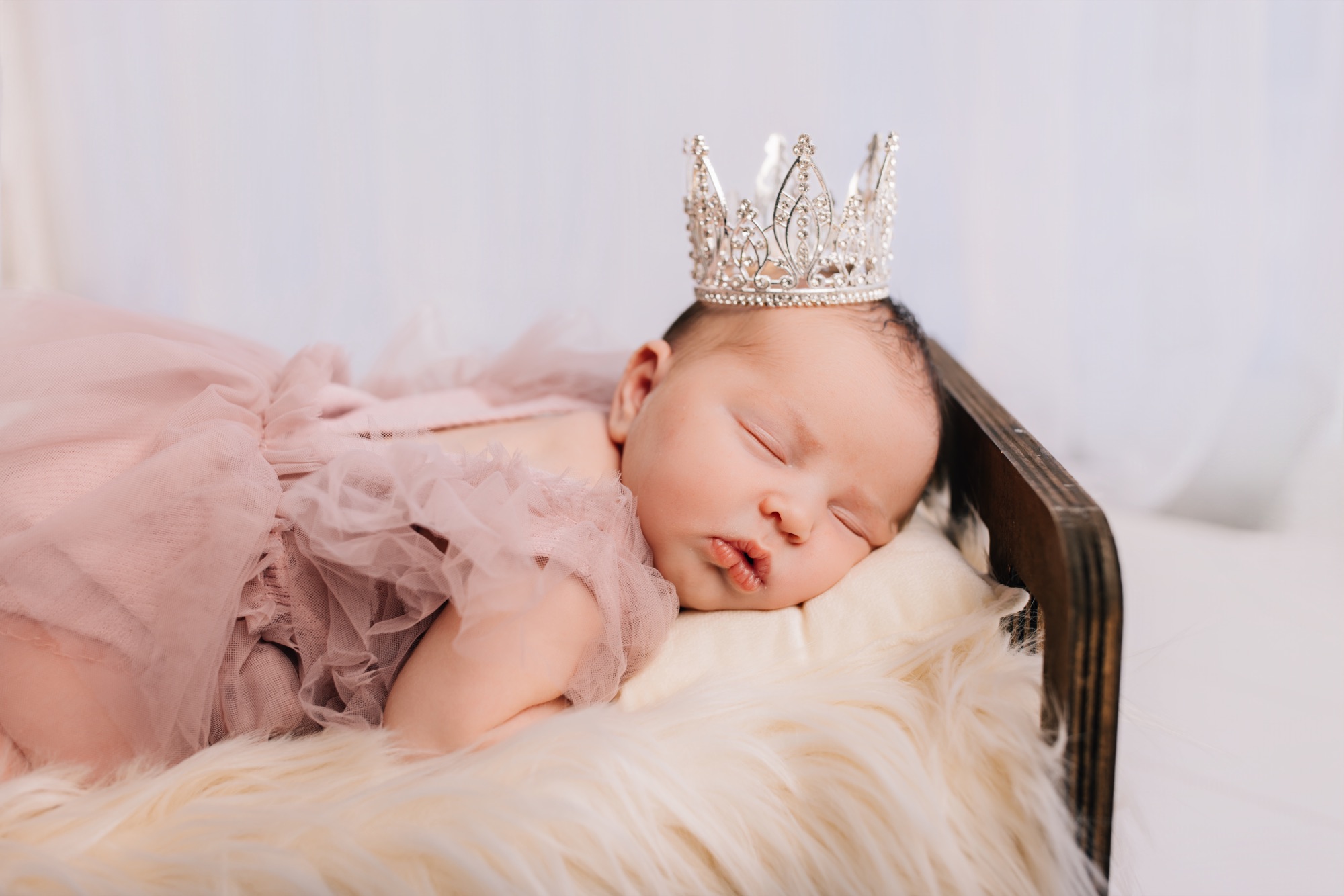 Sleeping newborn in a dusty pink tulle dress and rhinestone crown on a tiny wooden bed