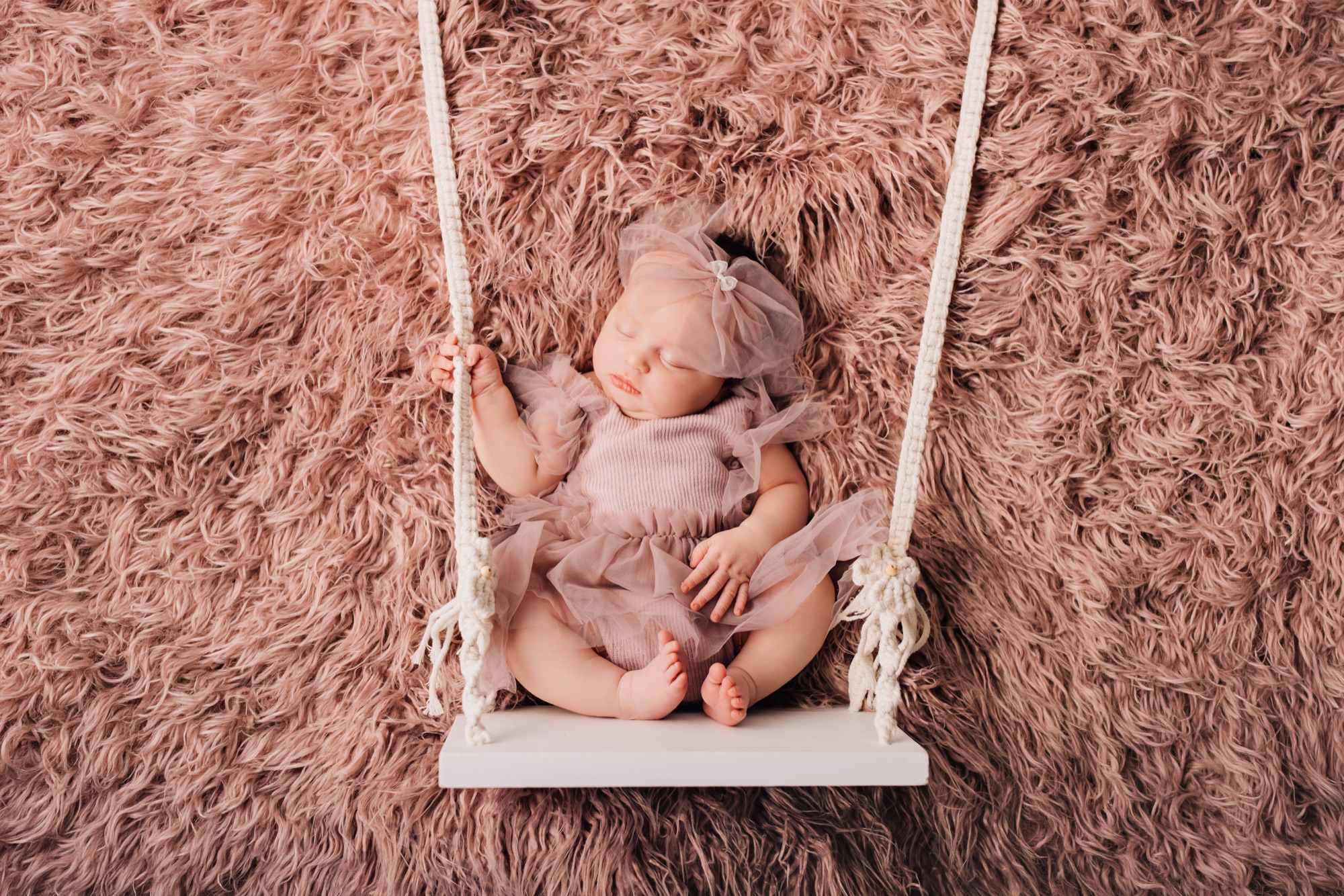 Sleeping newborn in a mauve tulle romper posed on a white swing prop, photographed from above