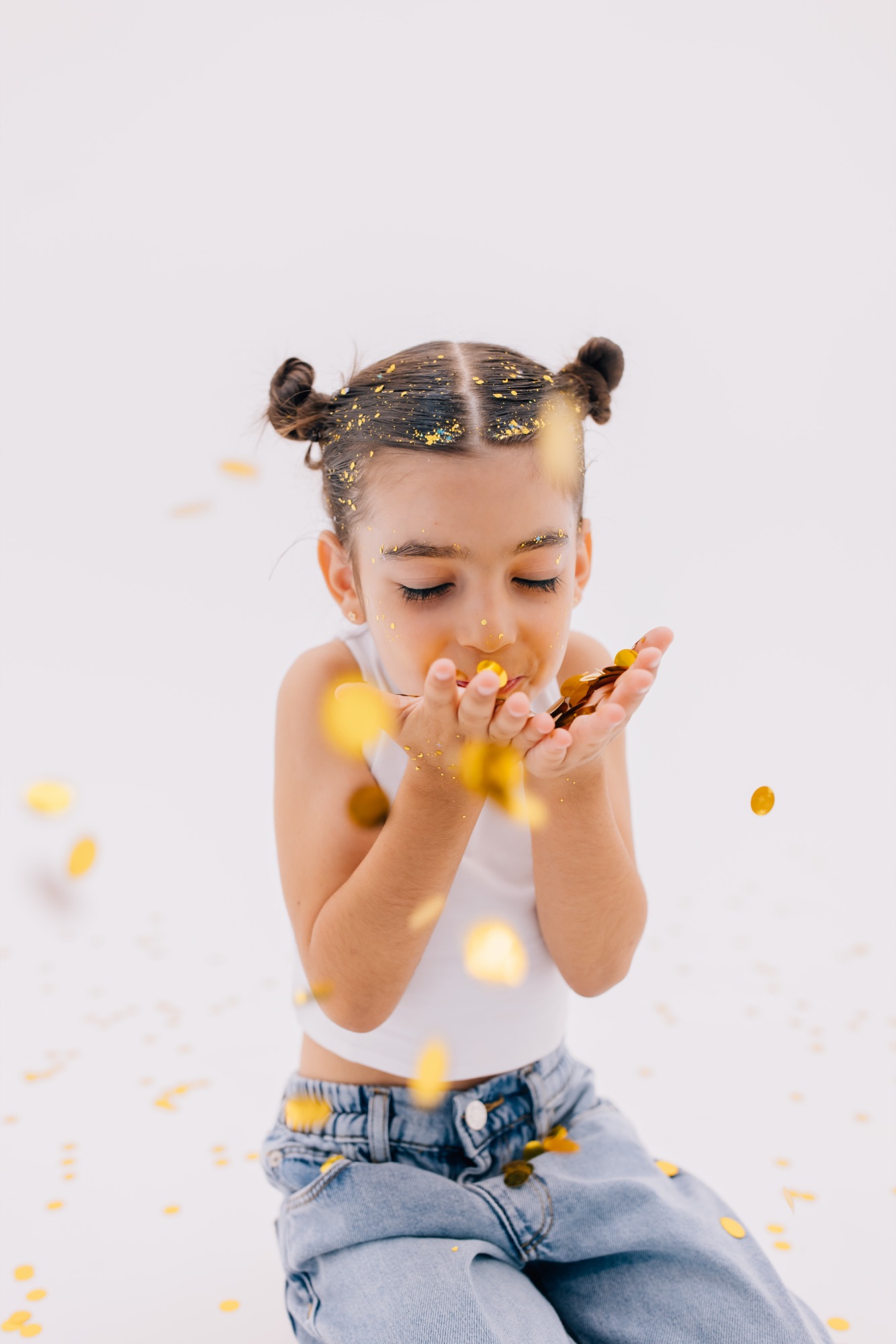 Girl with space buns blowing gold confetti from her hands in a white studio