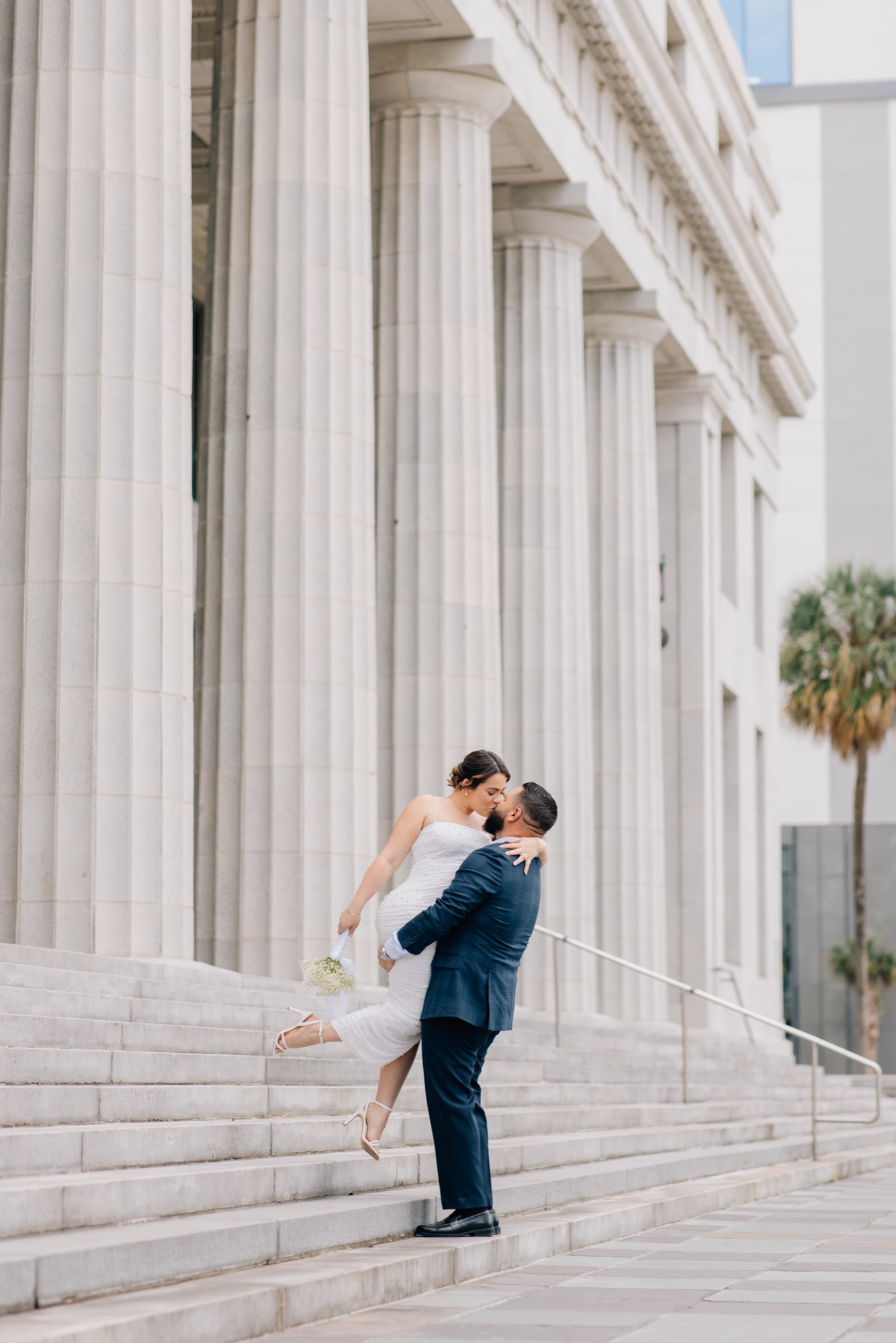 Groom in a navy suit lifting and kissing his bride on courthouse steps framed by stone columns and a palm tree
