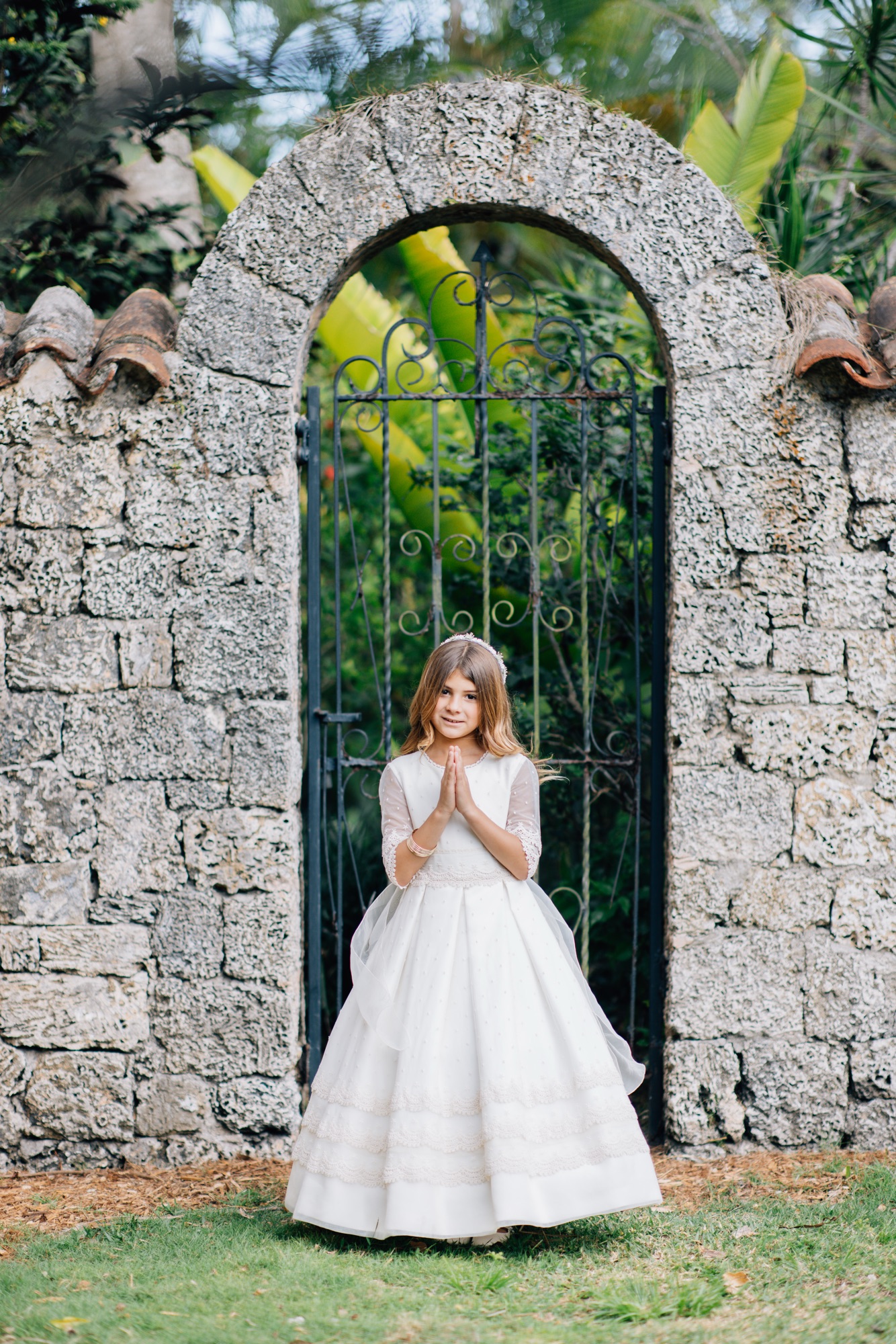Girl in a white first communion gown with hands folded in prayer beside a coral stone arch