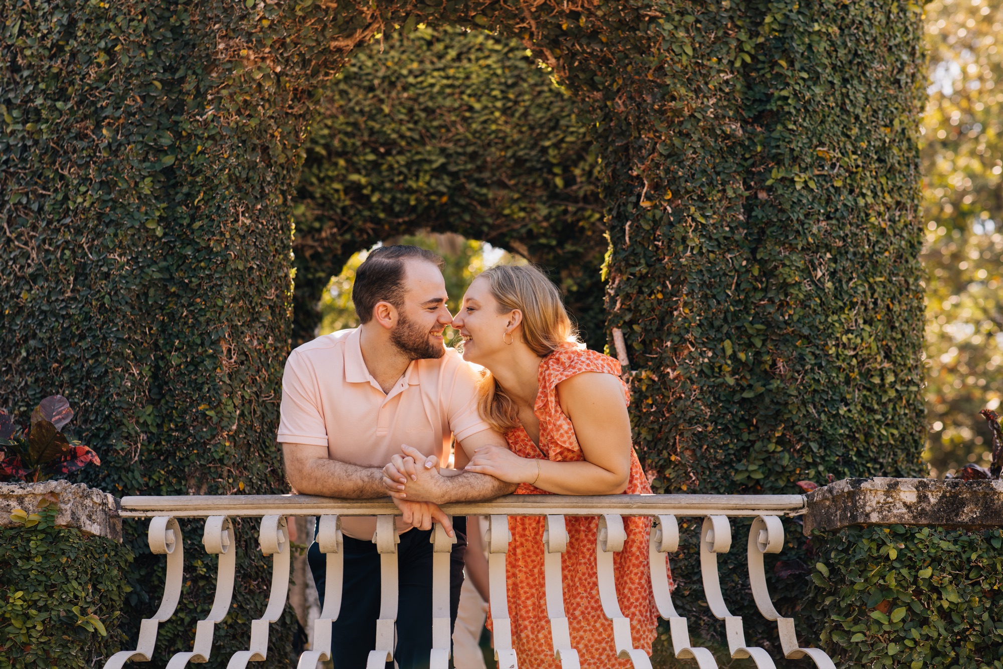 Engaged couple nose to nose on a white balustrade beneath ivy-covered garden arches at golden hour